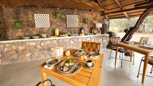 A stone wall with a thatched roof, a wooden table set with breakfast, and a stone bench.