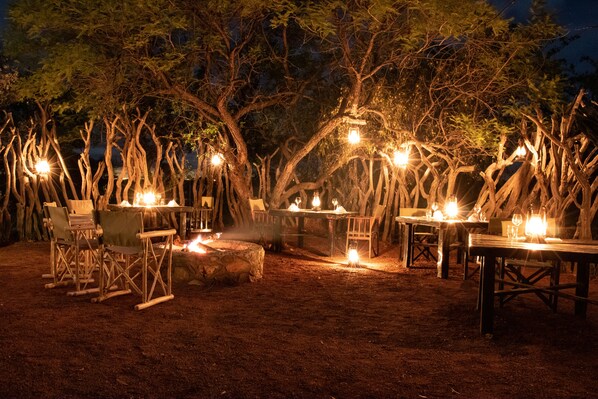 Un espacio de comedor al aire libre con mesas y sillas dispuestas bajo los árboles, rodeado por una valla de madera rústica.