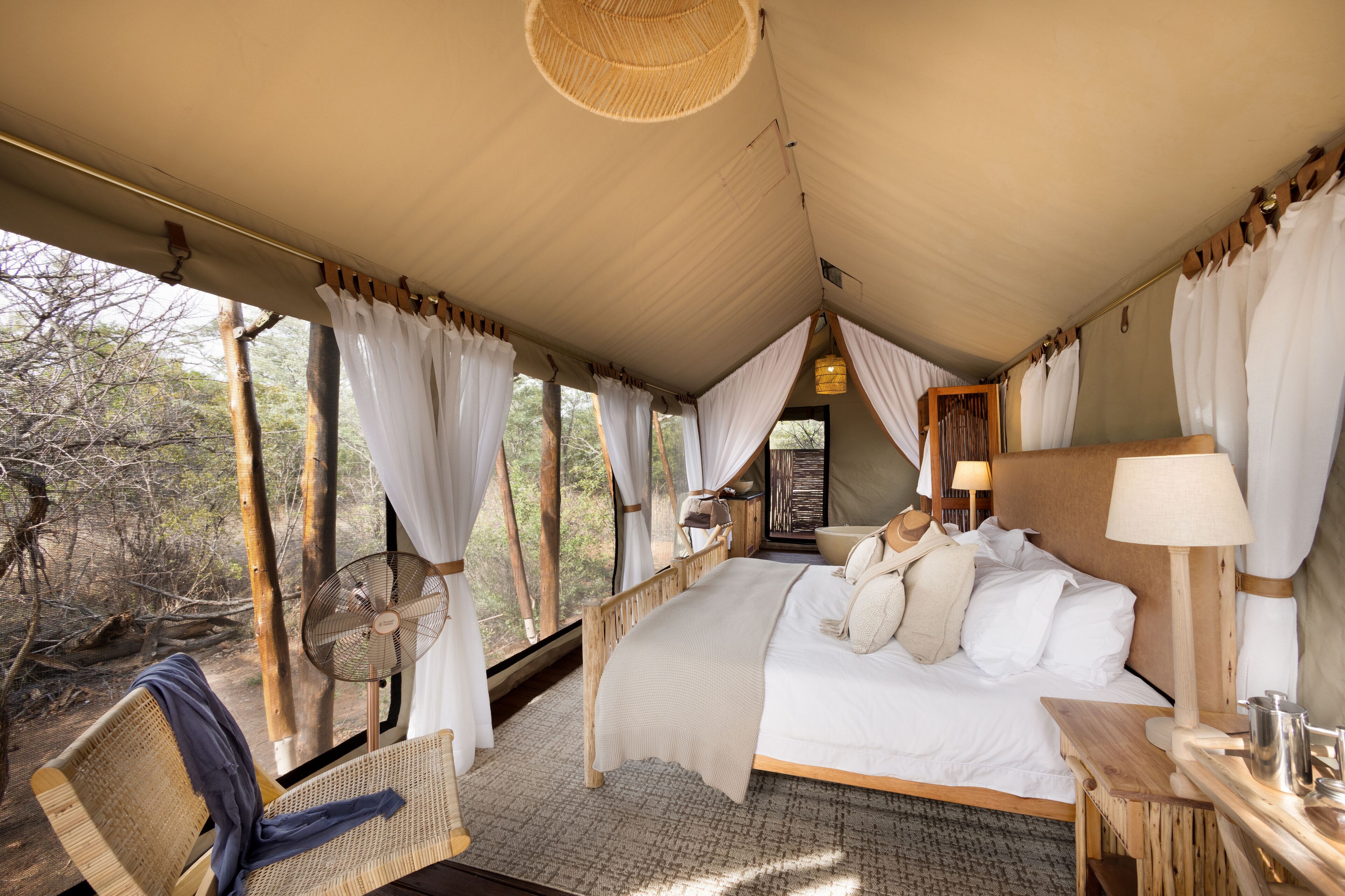 A spacious tented room with a large bed, a fan, and a view of the surrounding forest.
