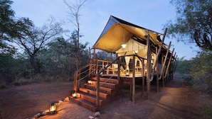 A rustic, elevated wooden structure with a large canopy and a staircase leading to a deck, surrounded by natural bushland.