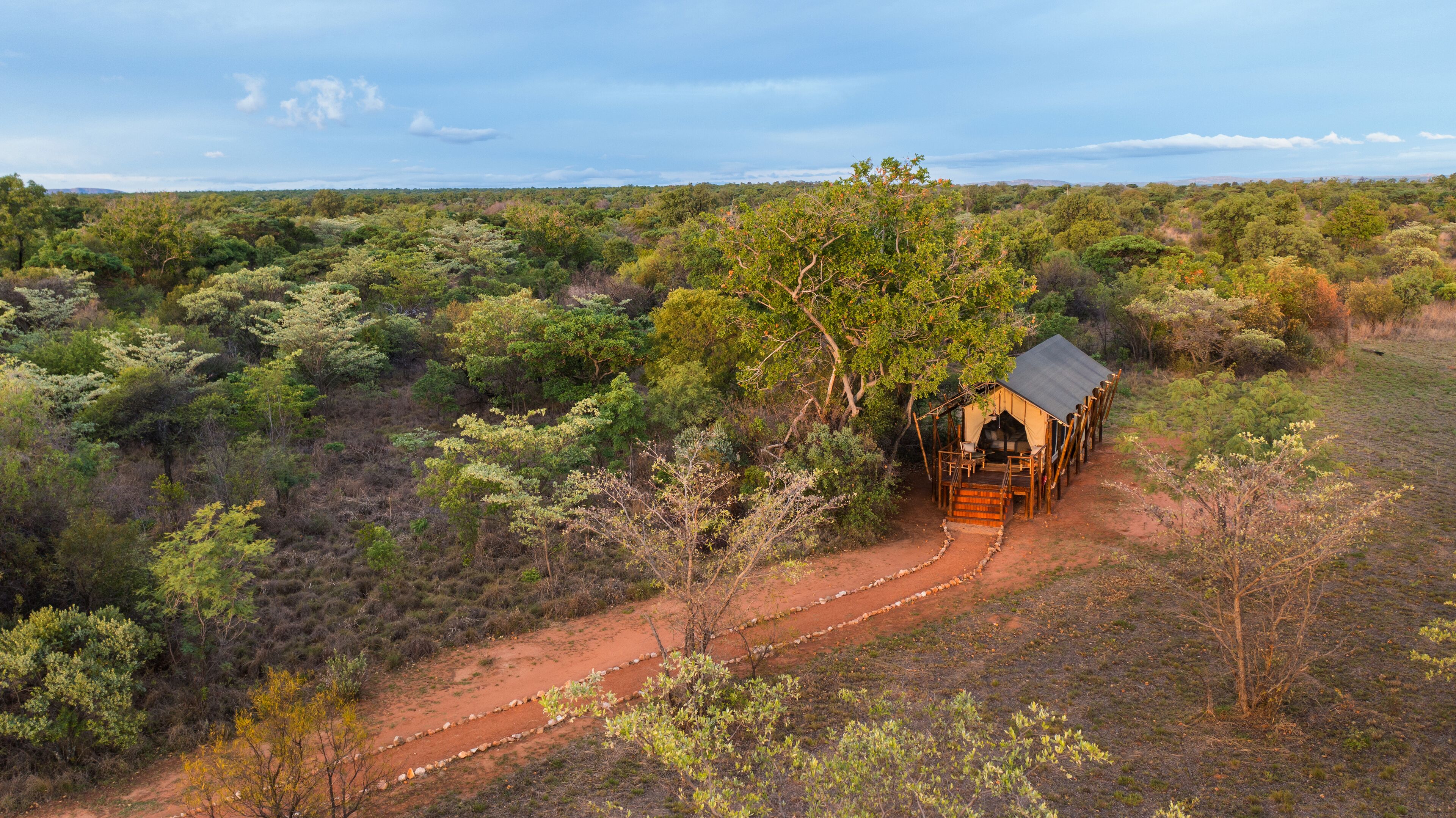 A safari lodge with a dirt path leading to it, surrounded by dense bushland.