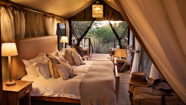 A tented accommodation with a bed, bedside lamps, a table, chairs, and a view of the outdoors.