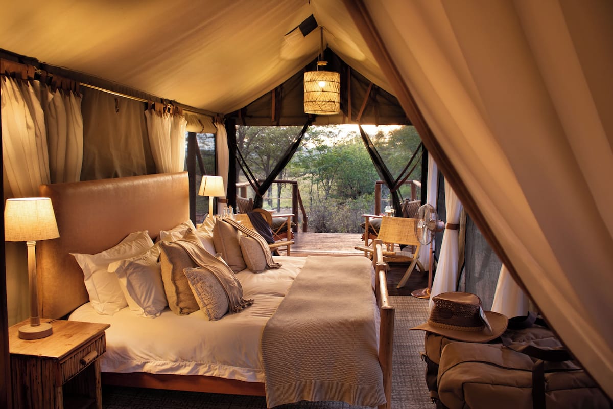 A tented accommodation with a bed, bedside lamps, a table, chairs, and a view of the outdoors.