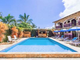 Piscina com espreguiçadeiras e guarda-sóis, cercada por palmeiras e um edifício de pedra com arcos.