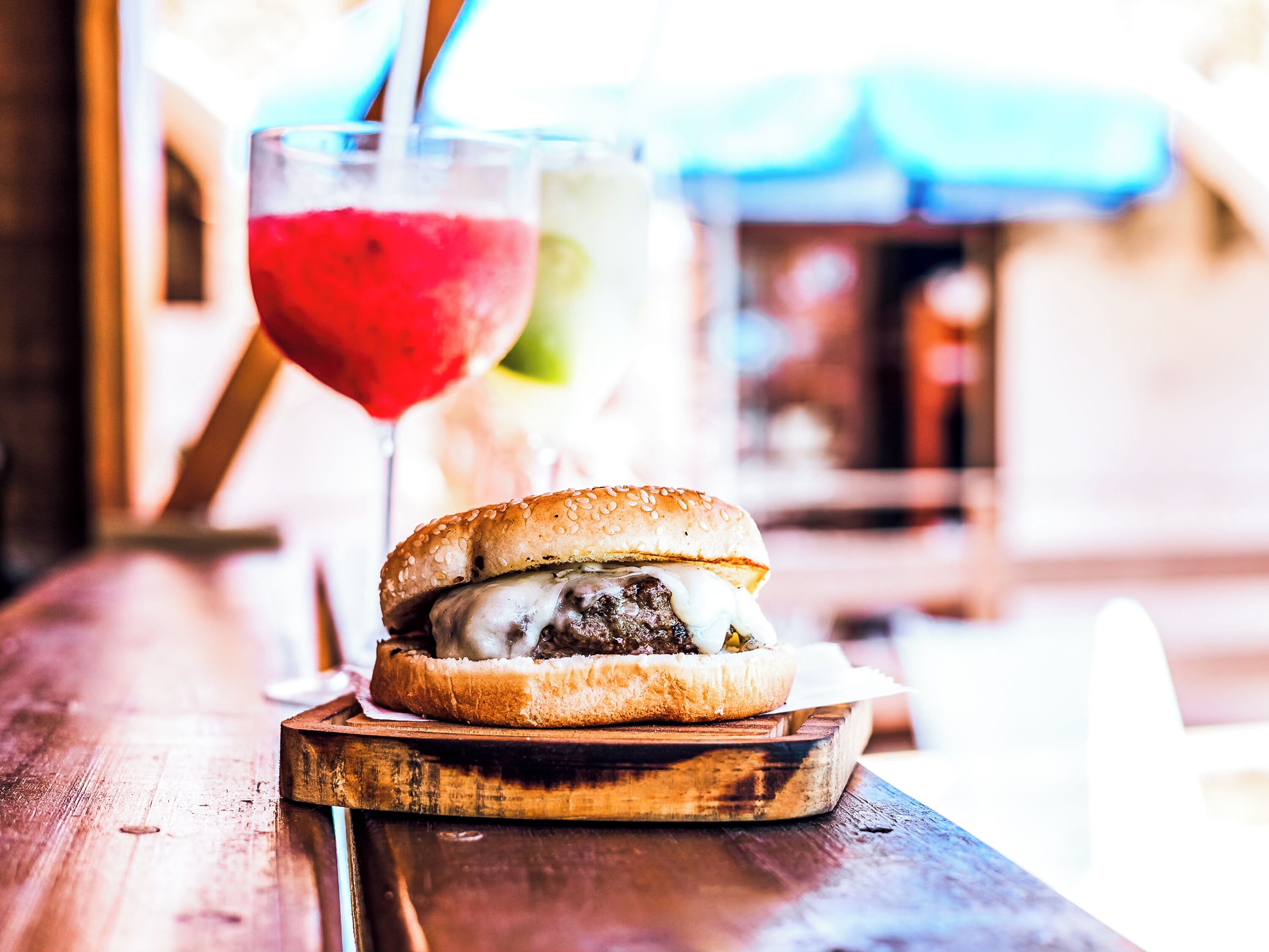 A burger on a wooden board with a drink in the background.