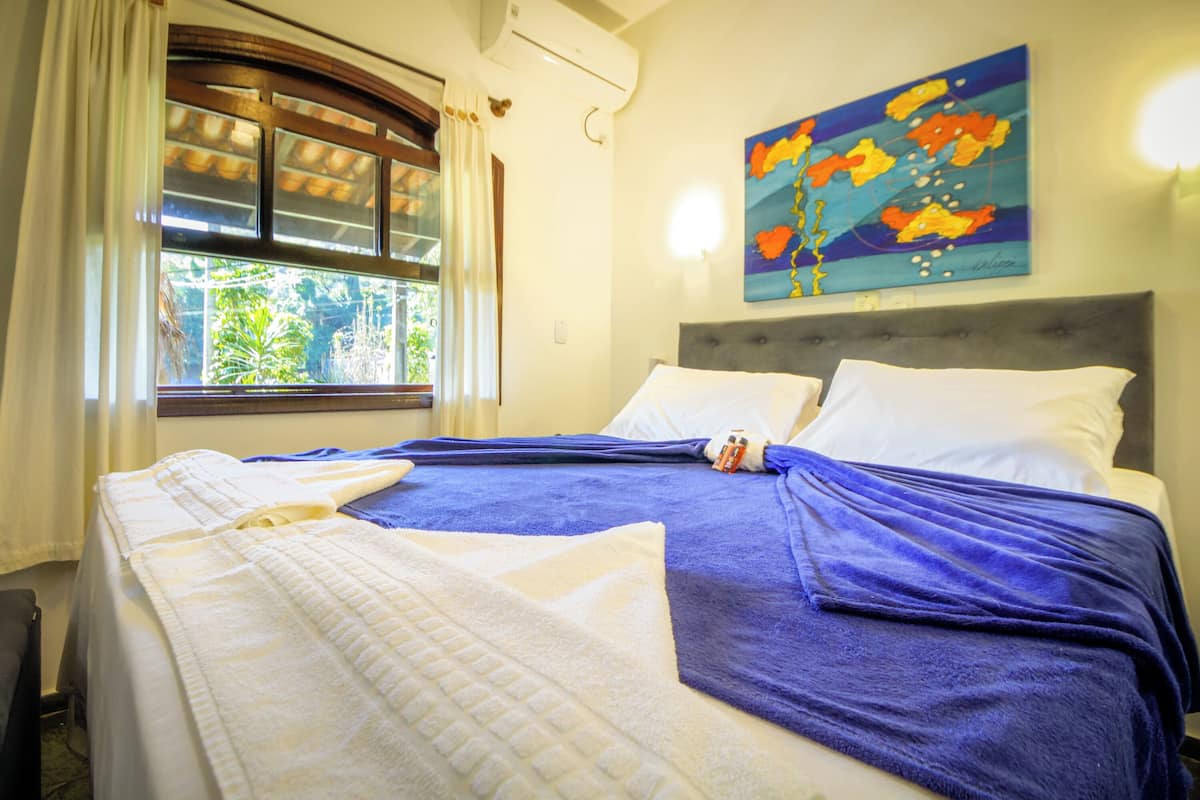 Une chambre d’hôtel avec un lit, une fenêtre avec des rideaux, un appareil de climatisation et un tableau coloré accroché au mur.