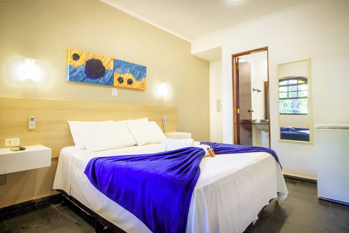 Une chambre d’hôtel avec un lit, deux tables de chevet, une commode, un tableau au mur et une porte donnant sur une autre pièce.