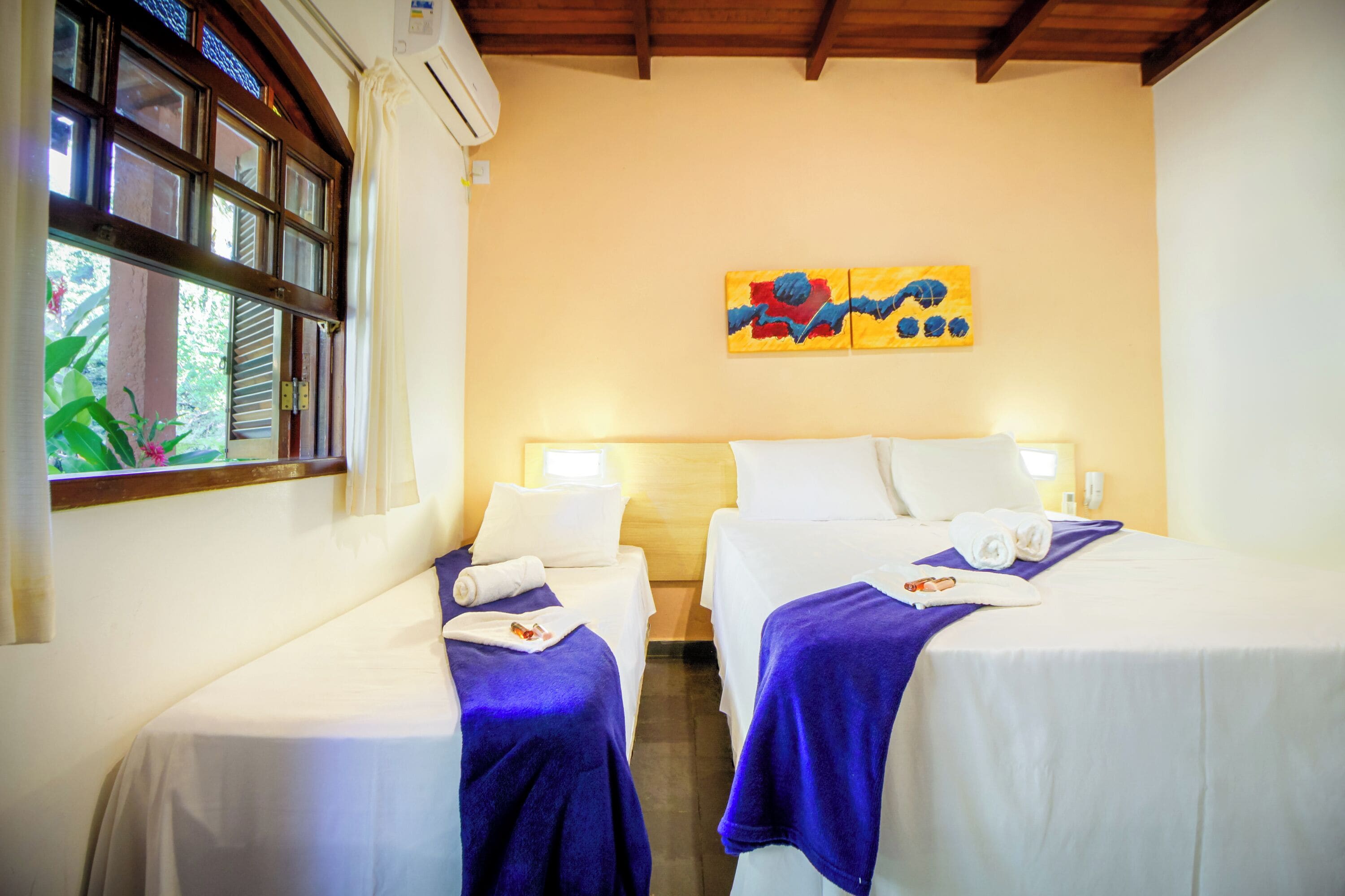 Een hotelkamer met twee bedden, een raam met zonwering, een schilderij aan de muur en een plafondventilator.