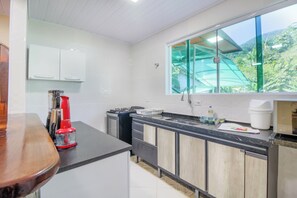 Enjoy cooking and sipping in this kitchen with a large window overlooking lush greenery, a stainless steel sink, and wooden cabinets.