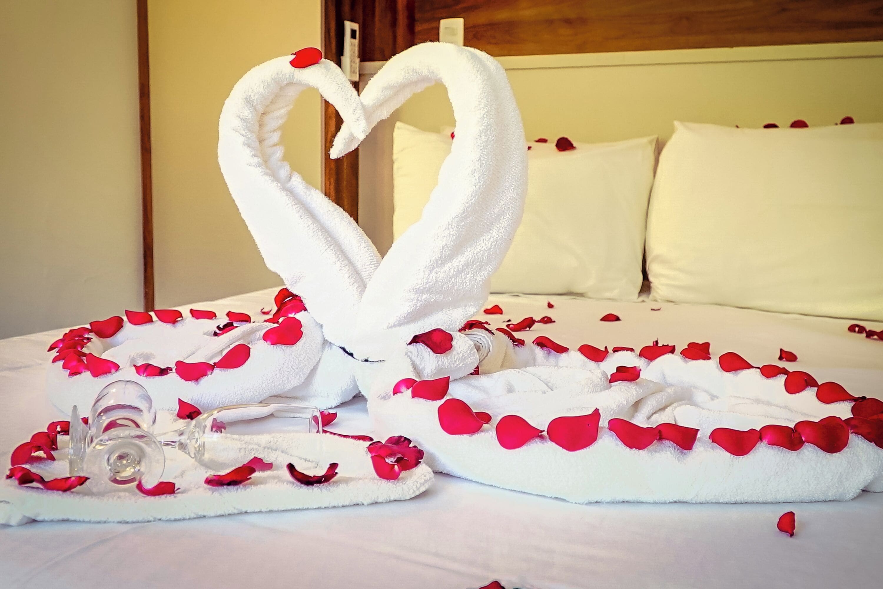 Una cama con sábanas blancas, una toalla dispuesta en forma de corazón y pétalos de rosa esparcidos.