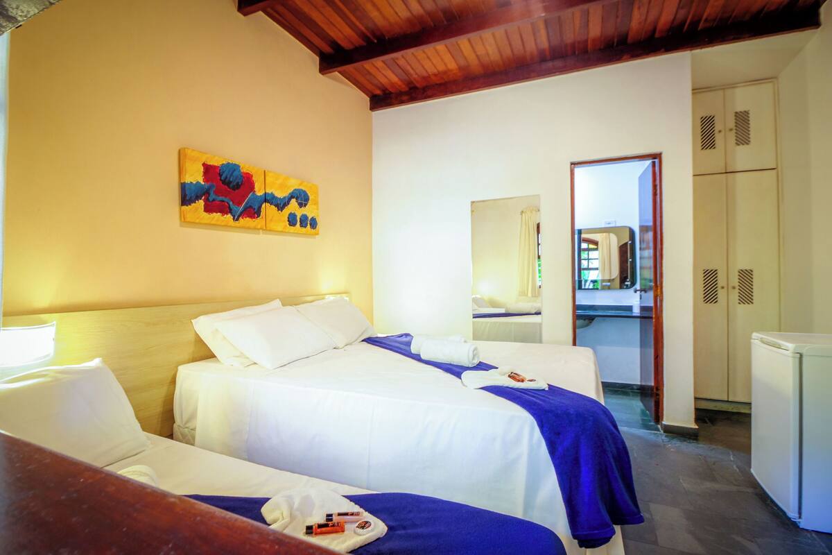 Une chambre d’hôtel avec deux lits, un tableau au mur, un miroir et, au fond, une petite cuisine.