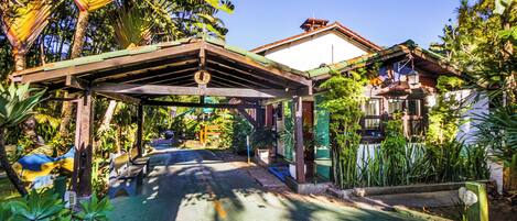 A covered walkway with benches, surrounded by lush greenery and a building with a red roof.