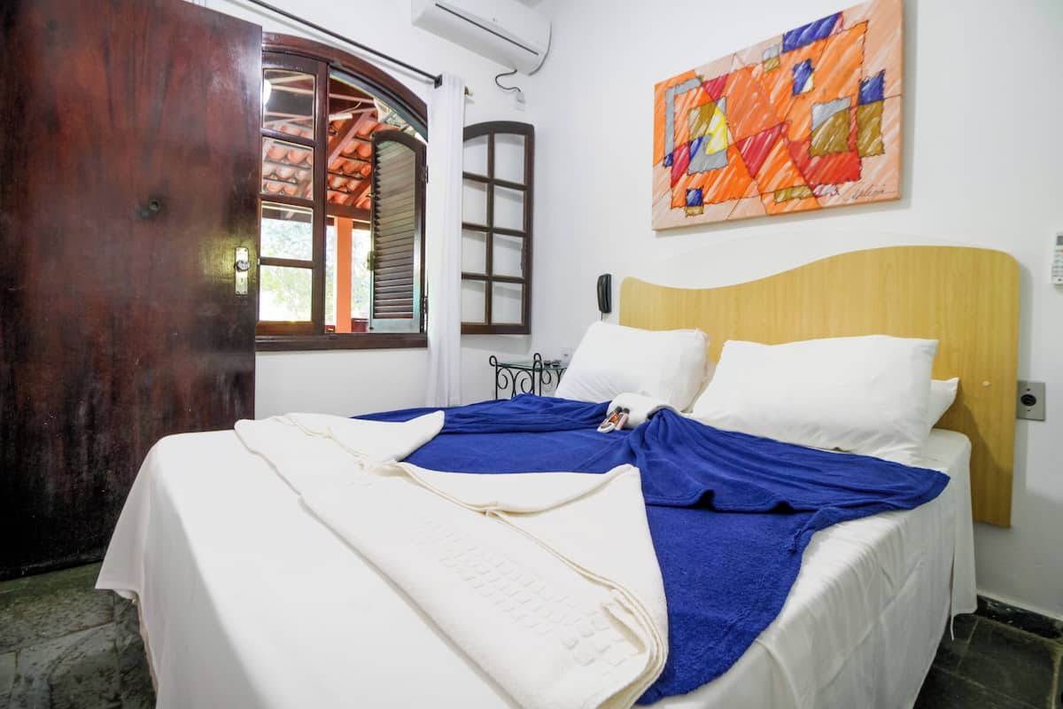 Une chambre à coucher avec un lit, une tête de lit en bois, un tableau abstrait aux couleurs vives et une fenêtre donnant sur un toit recouvert de tuiles.