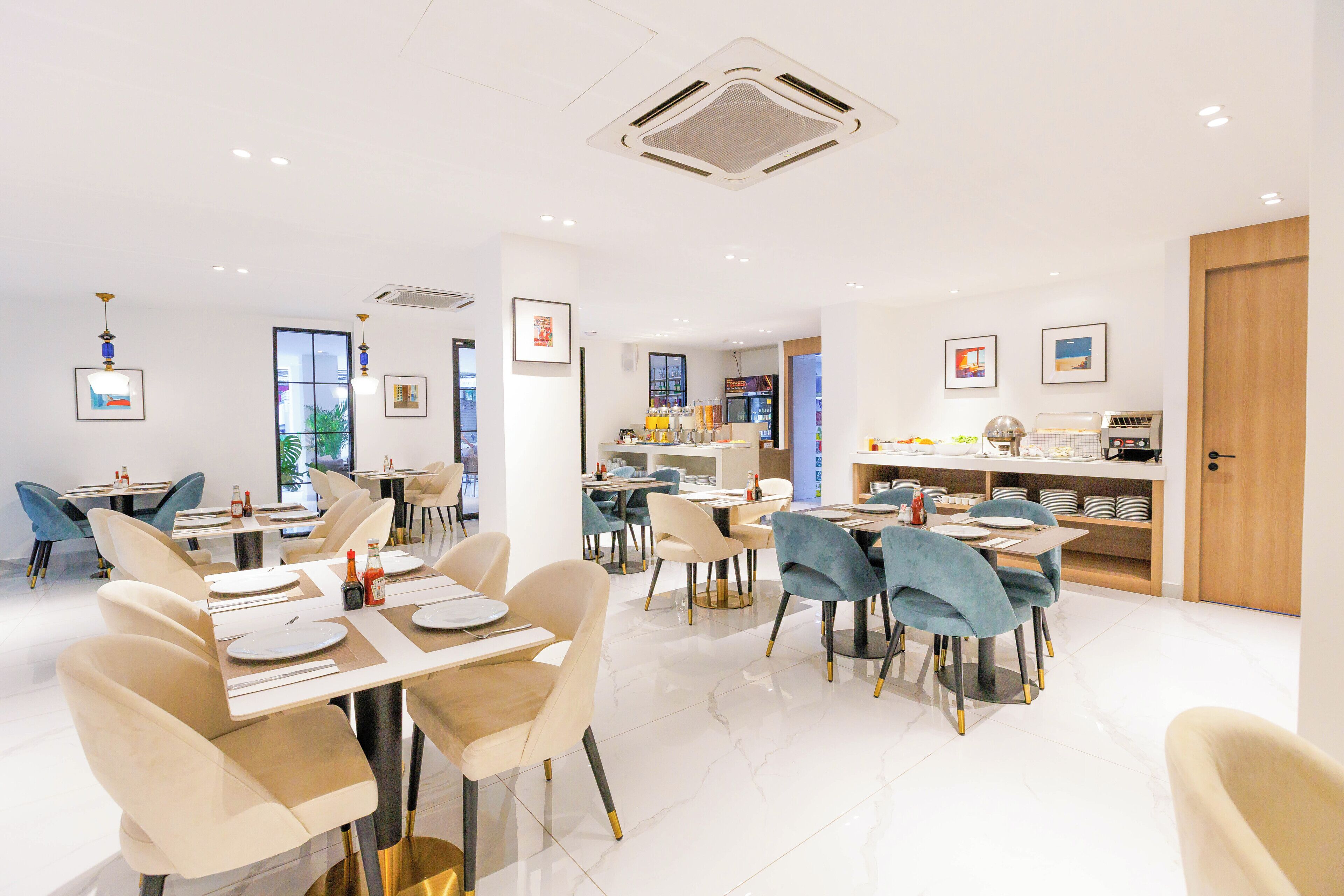 A modern dining area with multiple tables and chairs, a kitchen in the background, and framed pictures on the walls.