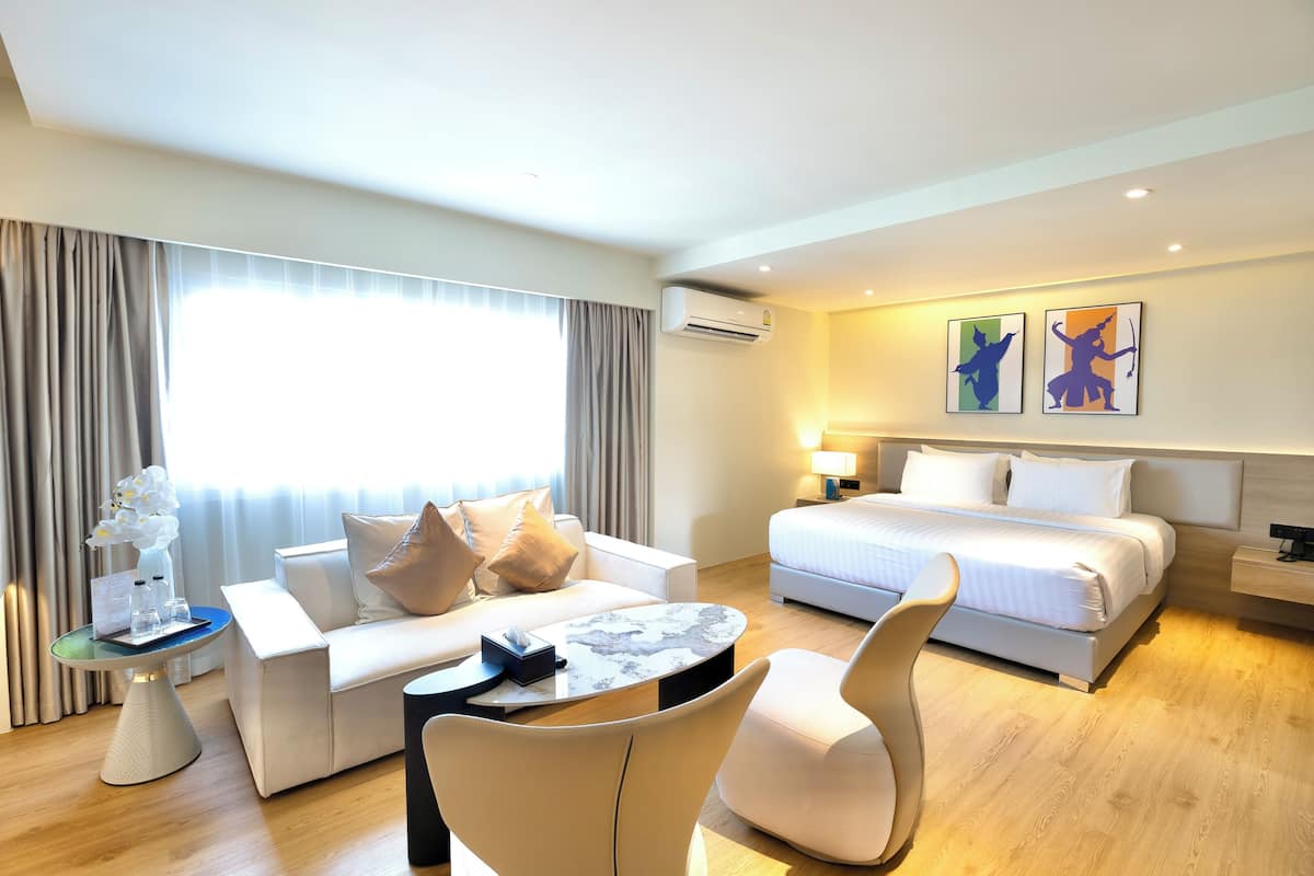 Une chambre d’hôtel avec un lit, un canapé, une table basse et une chaise. Il y a une fenêtre avec des rideaux, une lampe et deux œuvres d’art accrochées au mur.