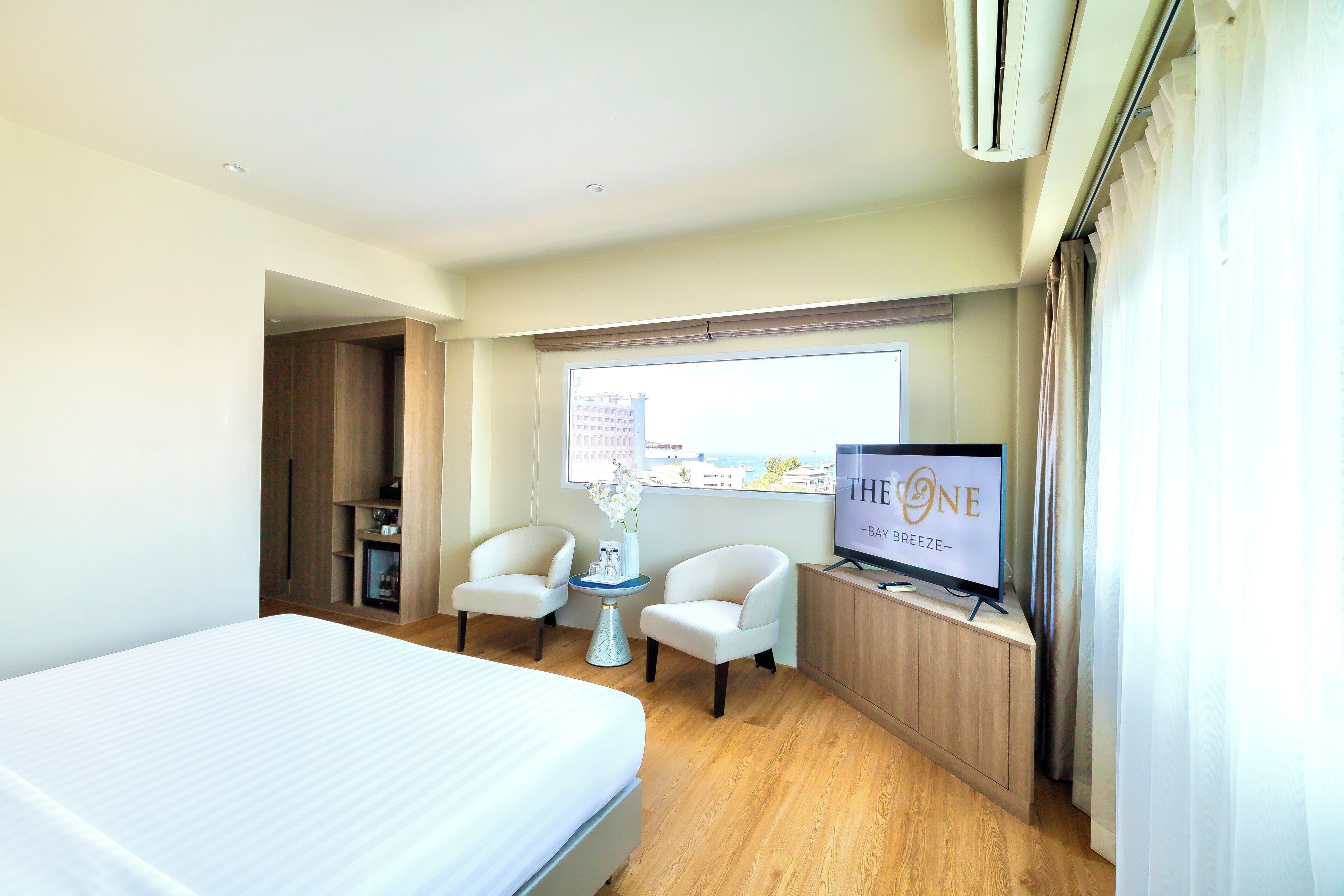 A hotel room with a bed, two armchairs, a small table, a TV, and a window with a city view.