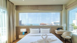 A hotel room with a large bed, a desk, a chair, and a view of the city through a large window. - The One Hotel Bay Breeze Pattaya (Pattaya)