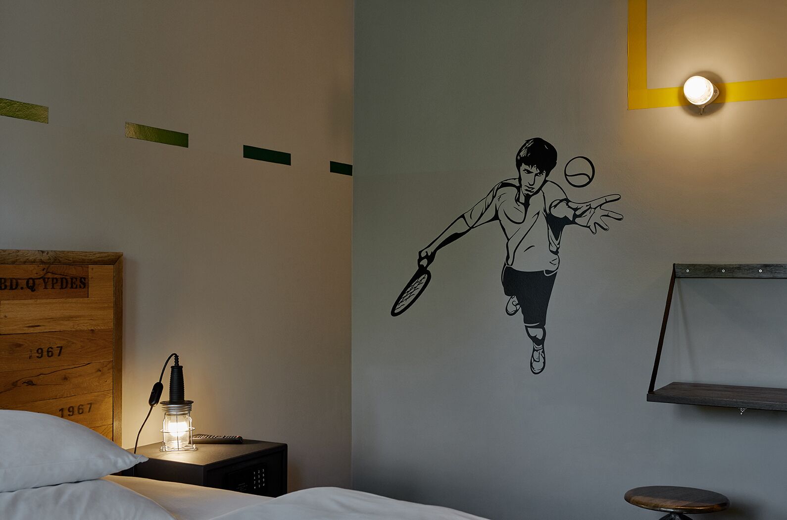 A bedroom with a bed, a nightstand with a lamp, a wall clock, and a wall-mounted shelf.