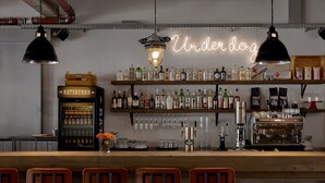 Un bar con un letrero de neón que dice "Underdog", estantes con botellas de licor, una máquina de café y taburetes de bar.