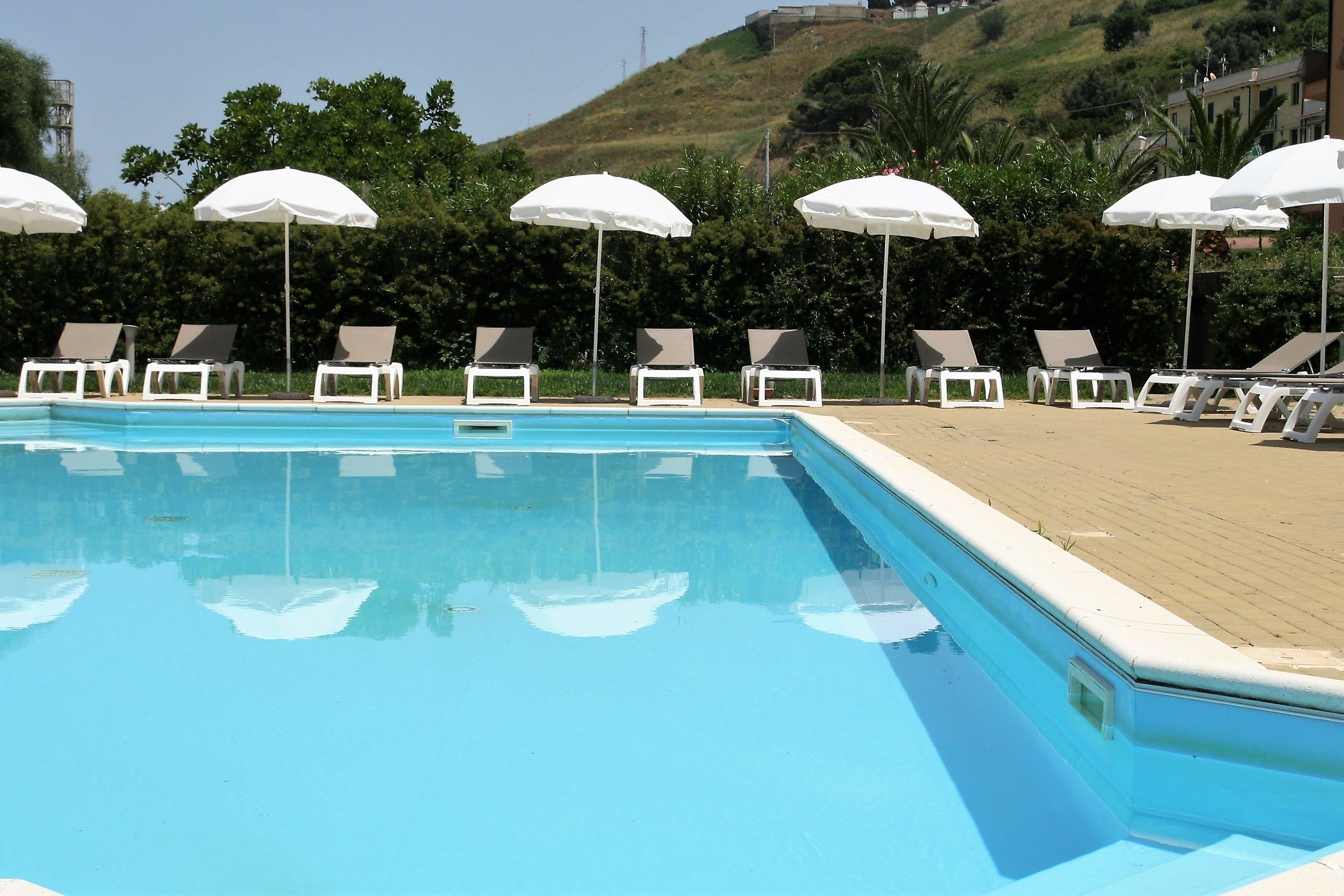 A swimming pool with white lounge chairs and umbrellas, surrounded by greenery and buildings in the background.