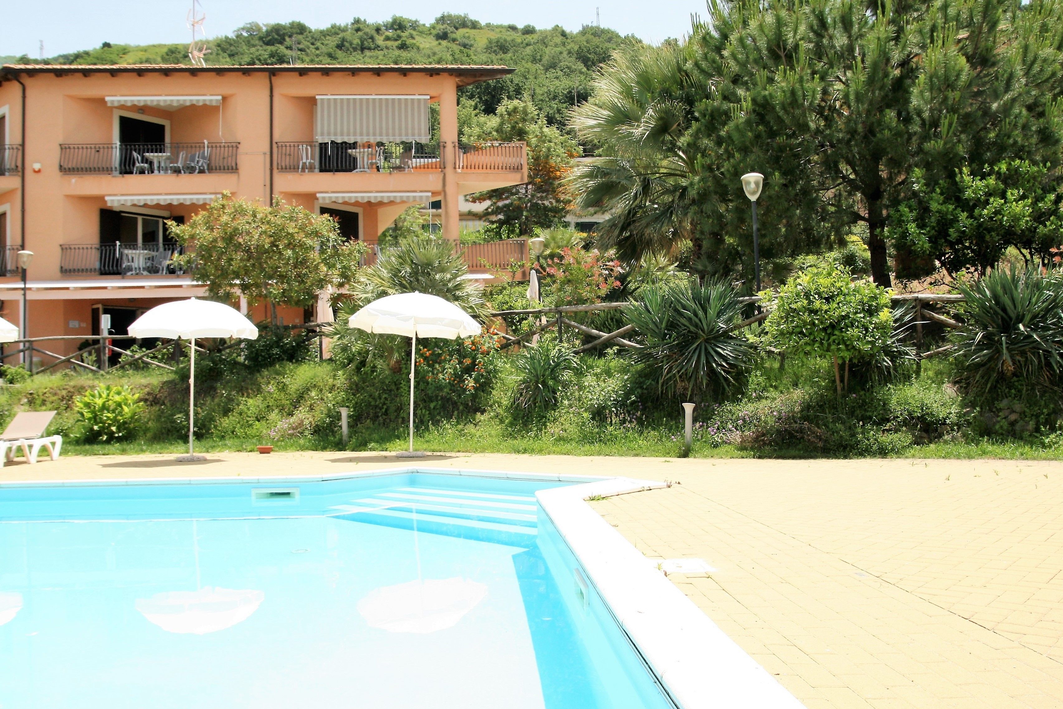 Una piscina con lettini e ombrelloni, circondata da vegetazione rigogliosa e un edificio a più piani con balconi.