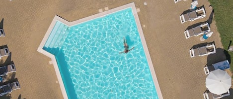 An aerial view of a rectangular swimming pool surrounded by lounge chairs and umbrellas, with a paved walkway around it.