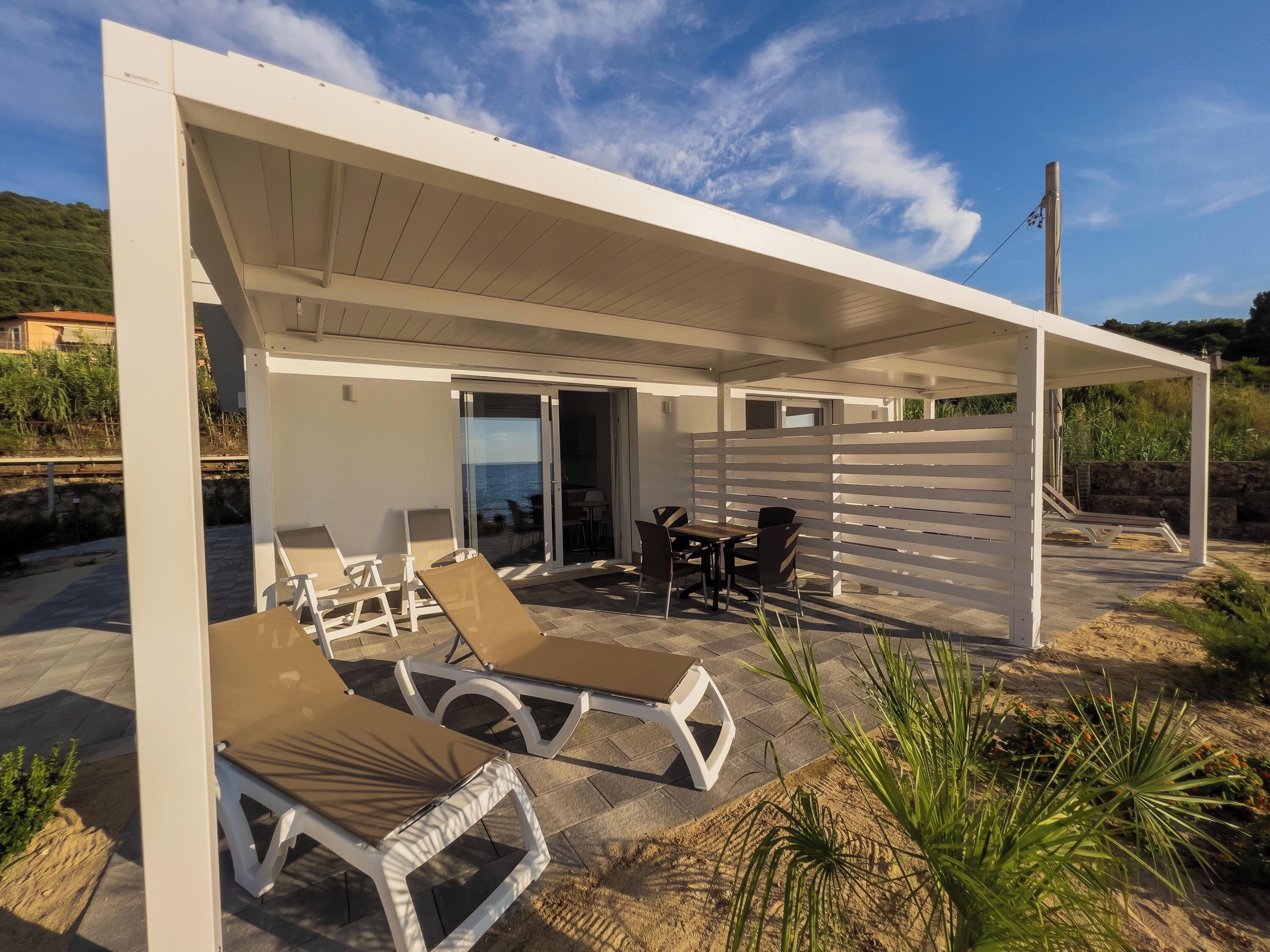 A covered outdoor area with lounge chairs, a table, and a view of the sea.