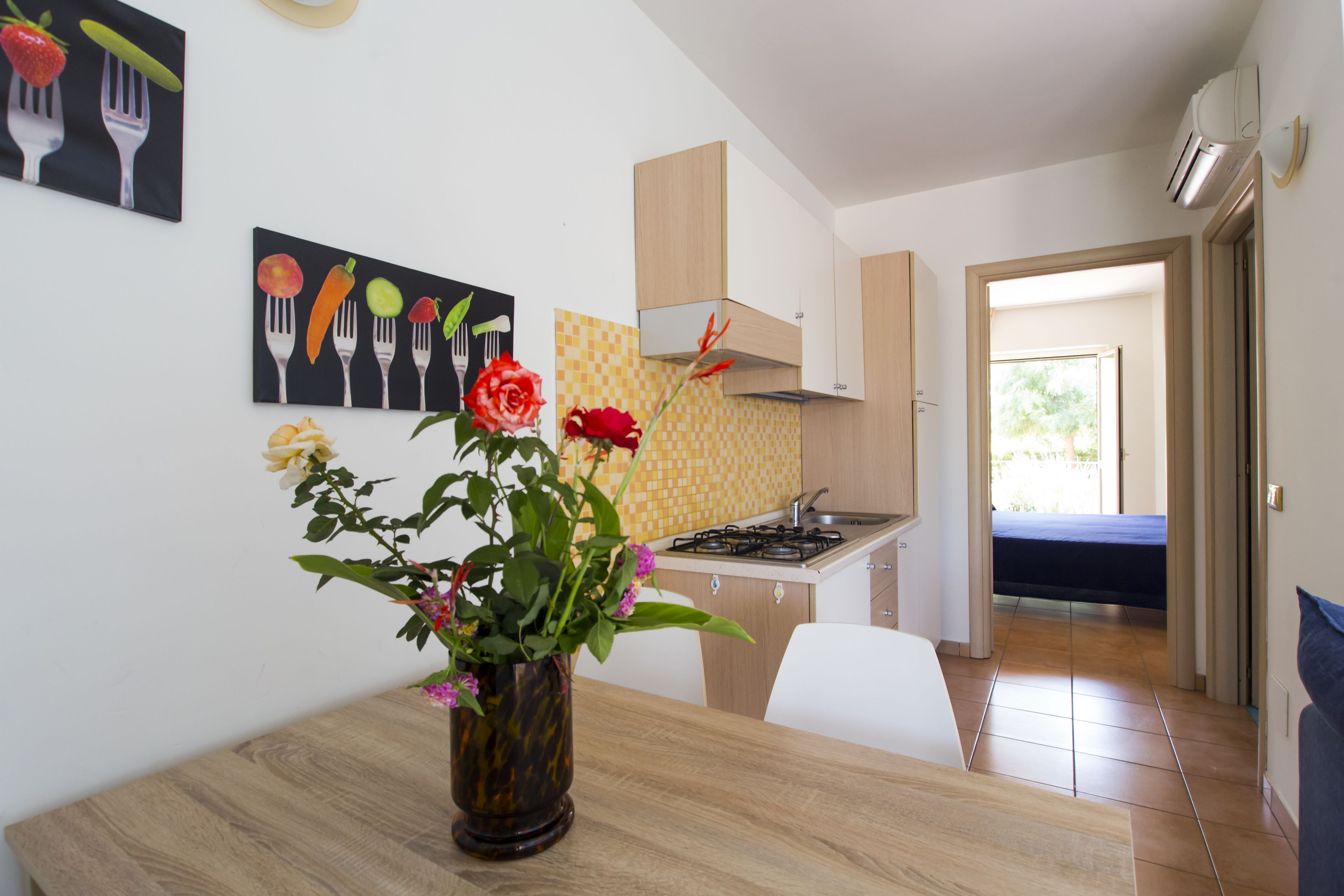 A modern kitchen with a dining table, a vase with flowers, and a painting of fruits and utensils on the wall.