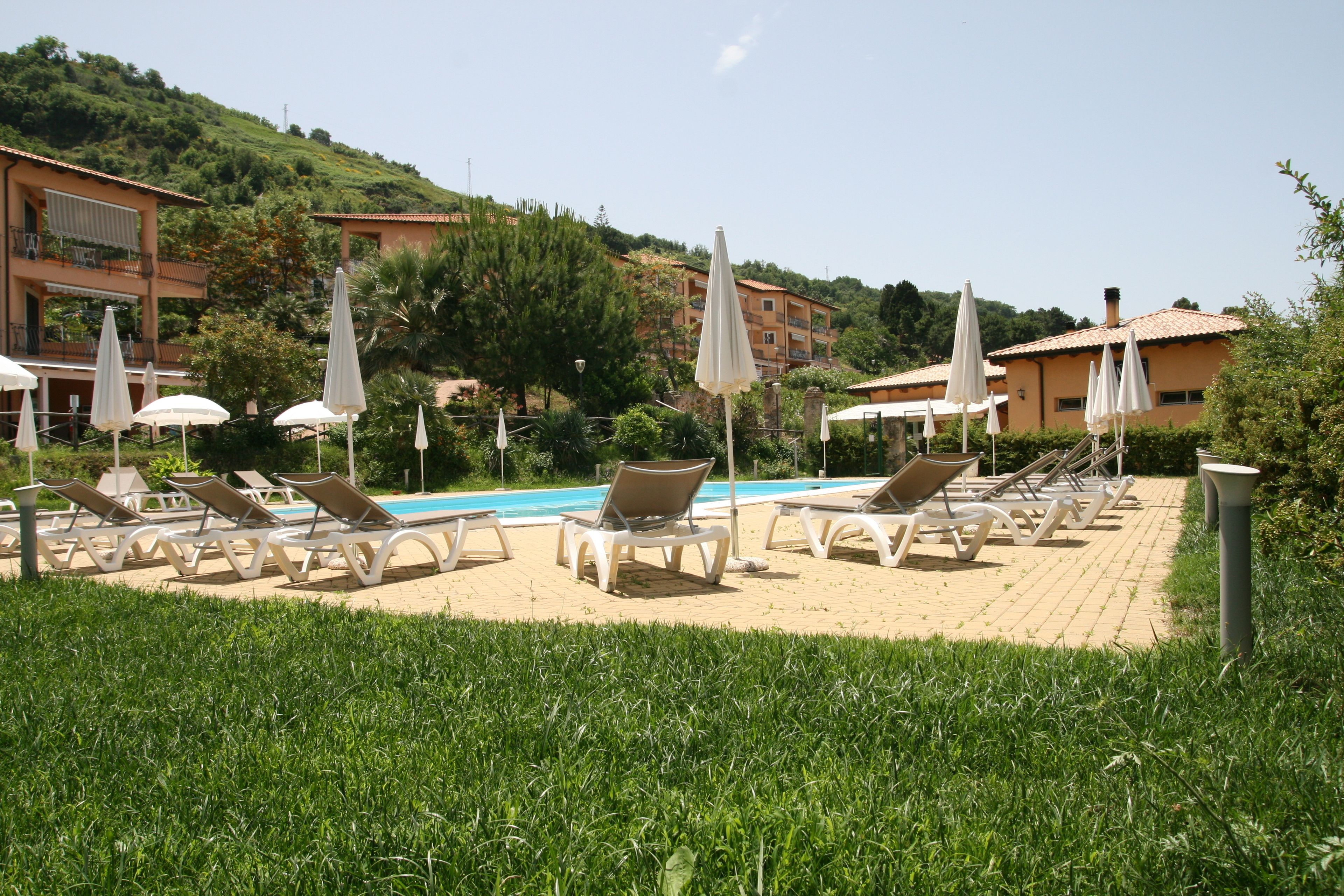 A poolside area with lounge chairs and umbrellas, surrounded by greenery and buildings in the background.
