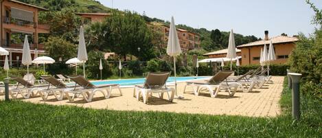 A poolside area with lounge chairs and umbrellas, surrounded by greenery and buildings in the background.