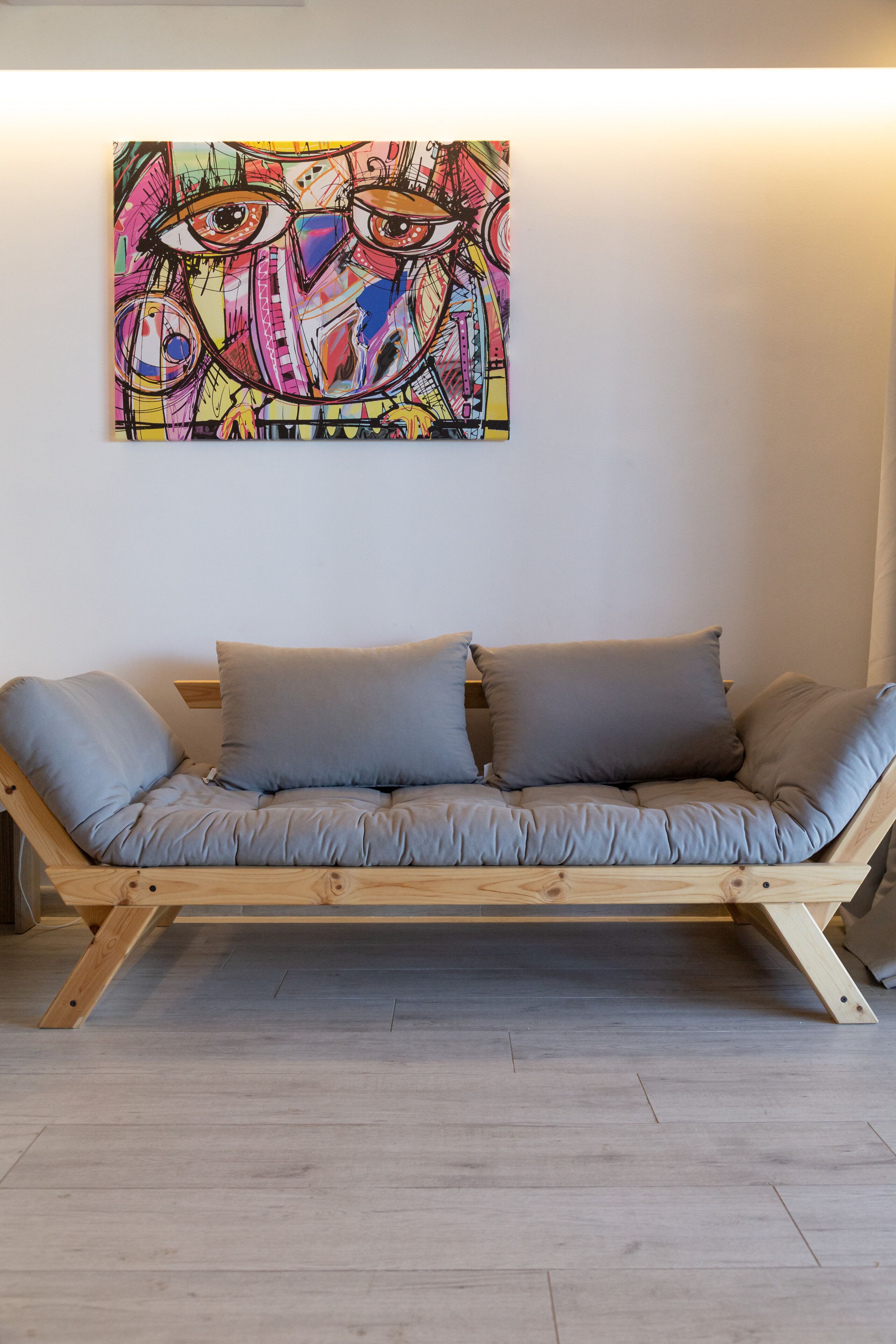 A wooden sofa with grey cushions against a white wall, with a colorful abstract painting above it.