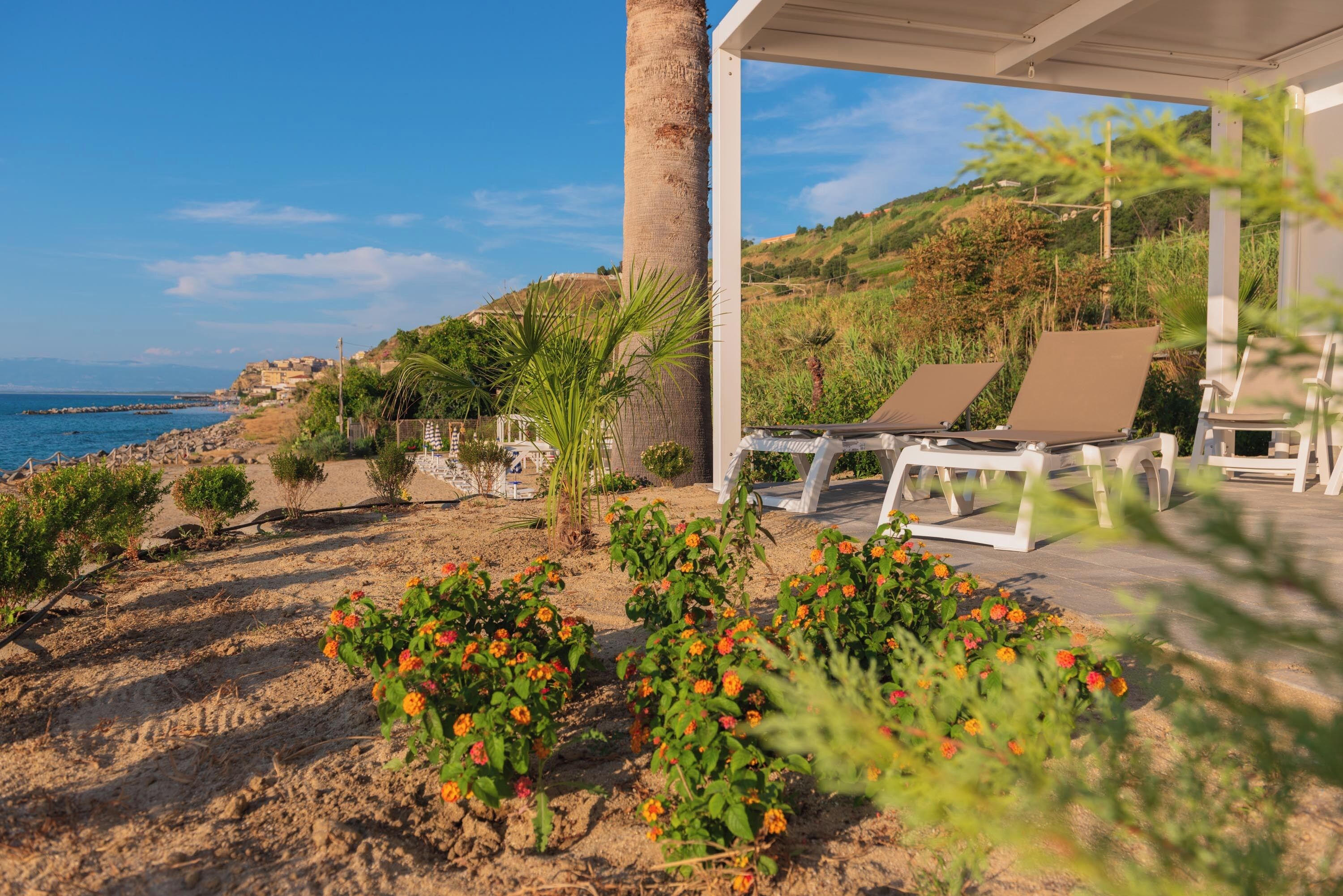 A covered outdoor seating area with two lounge chairs, a palm tree, and a view of the sea and hills.