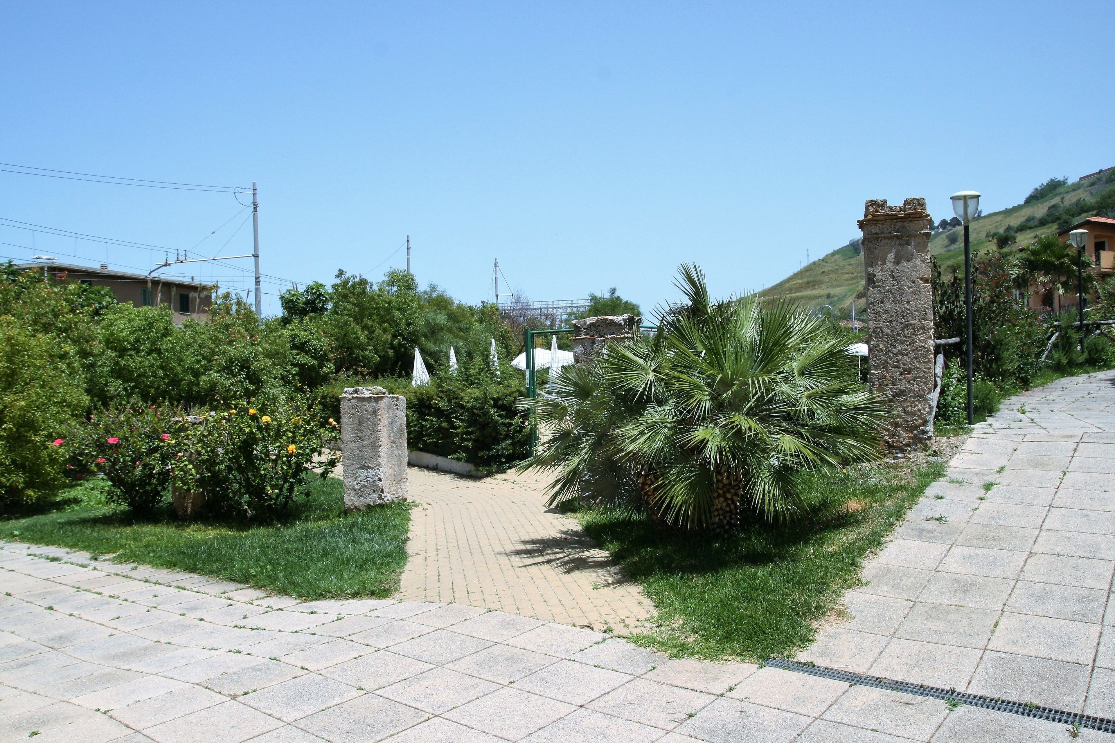 Un vialetto lastricato che conduce all'ingresso di un edificio, circondato da vegetazione e un pilastro in pietra.