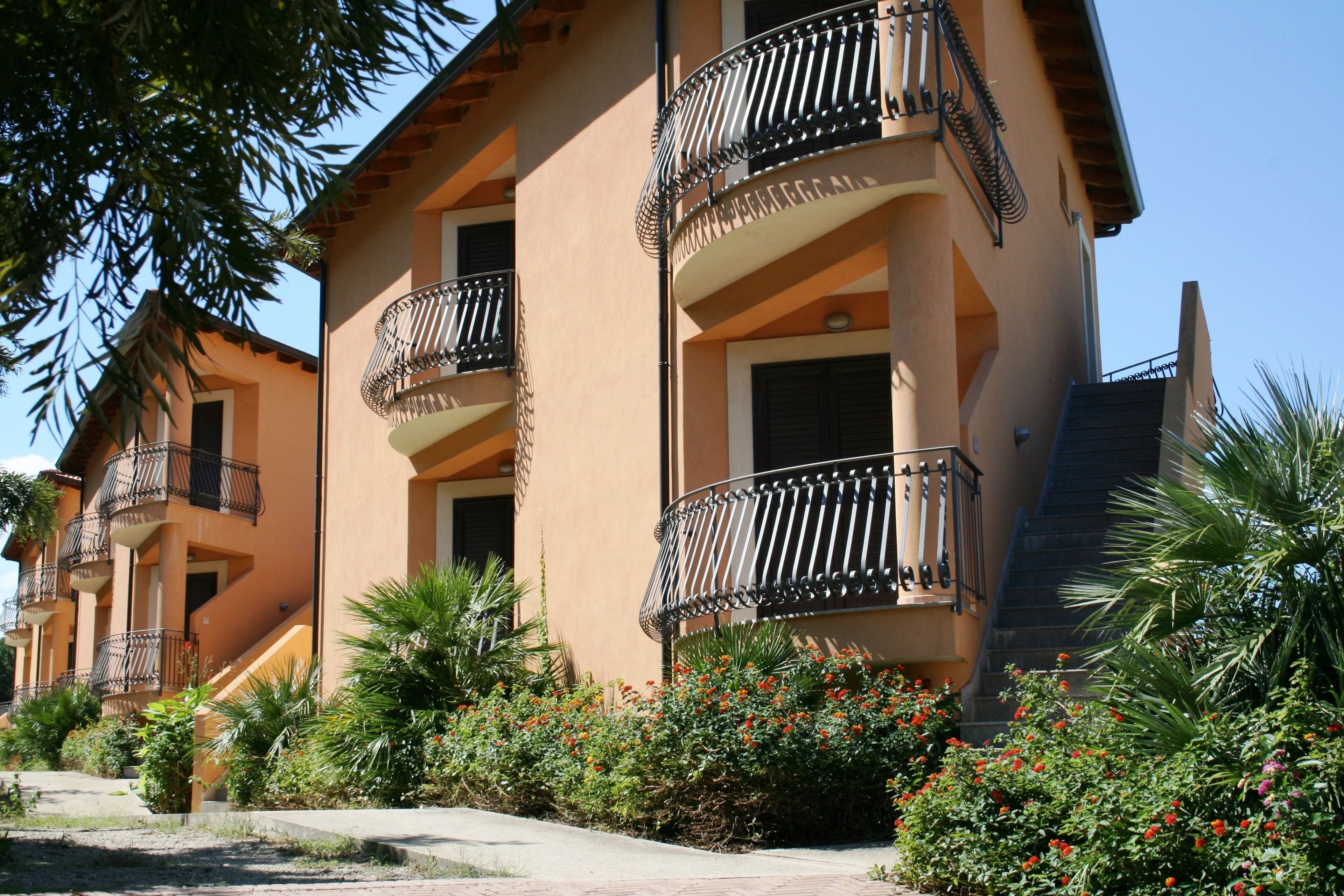 A multi-story building with balconies, surrounded by greenery and flowers.