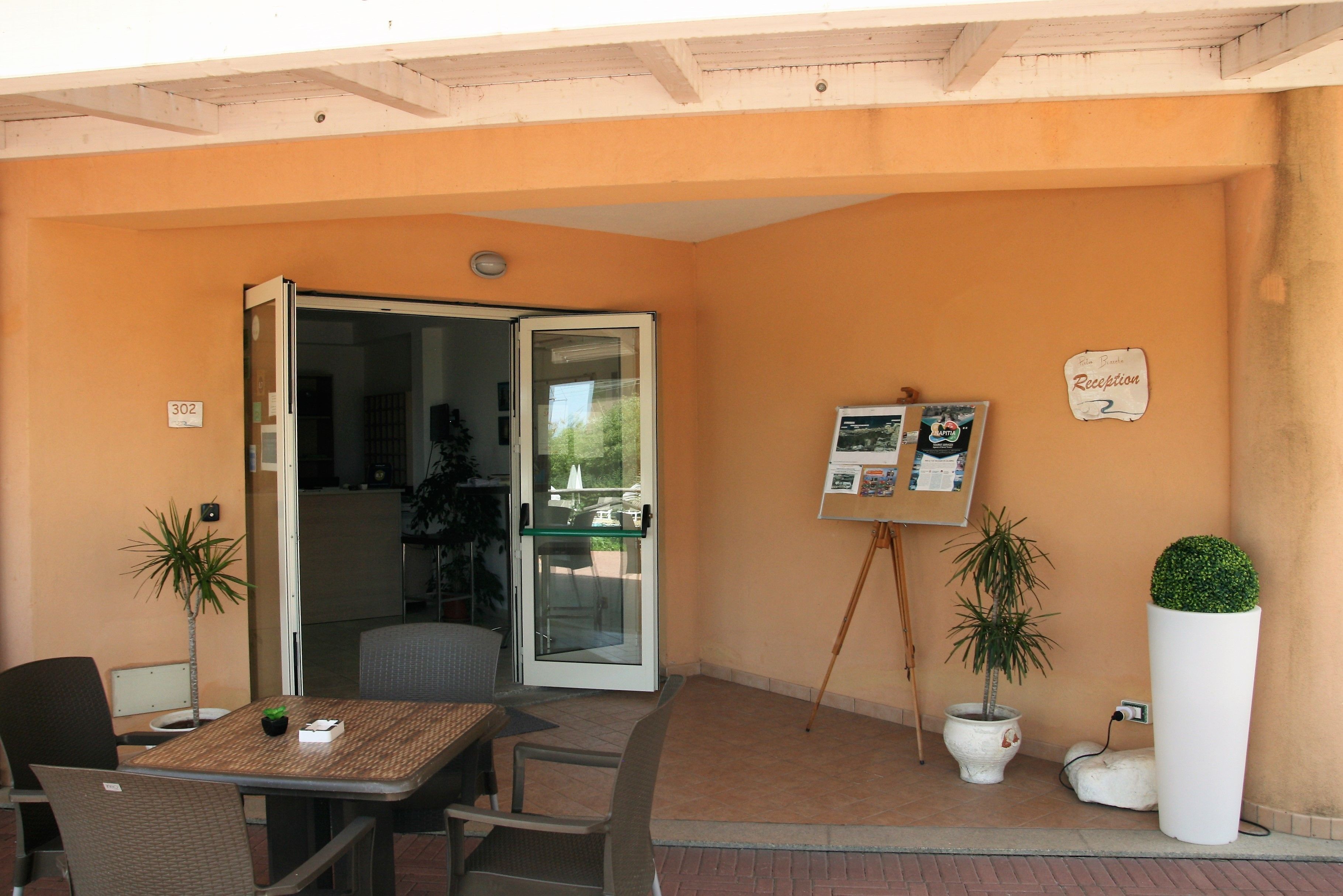 Un espace extérieur aménagé avec une table et des chaises, un panneau d’accueil et deux plantes en pot.