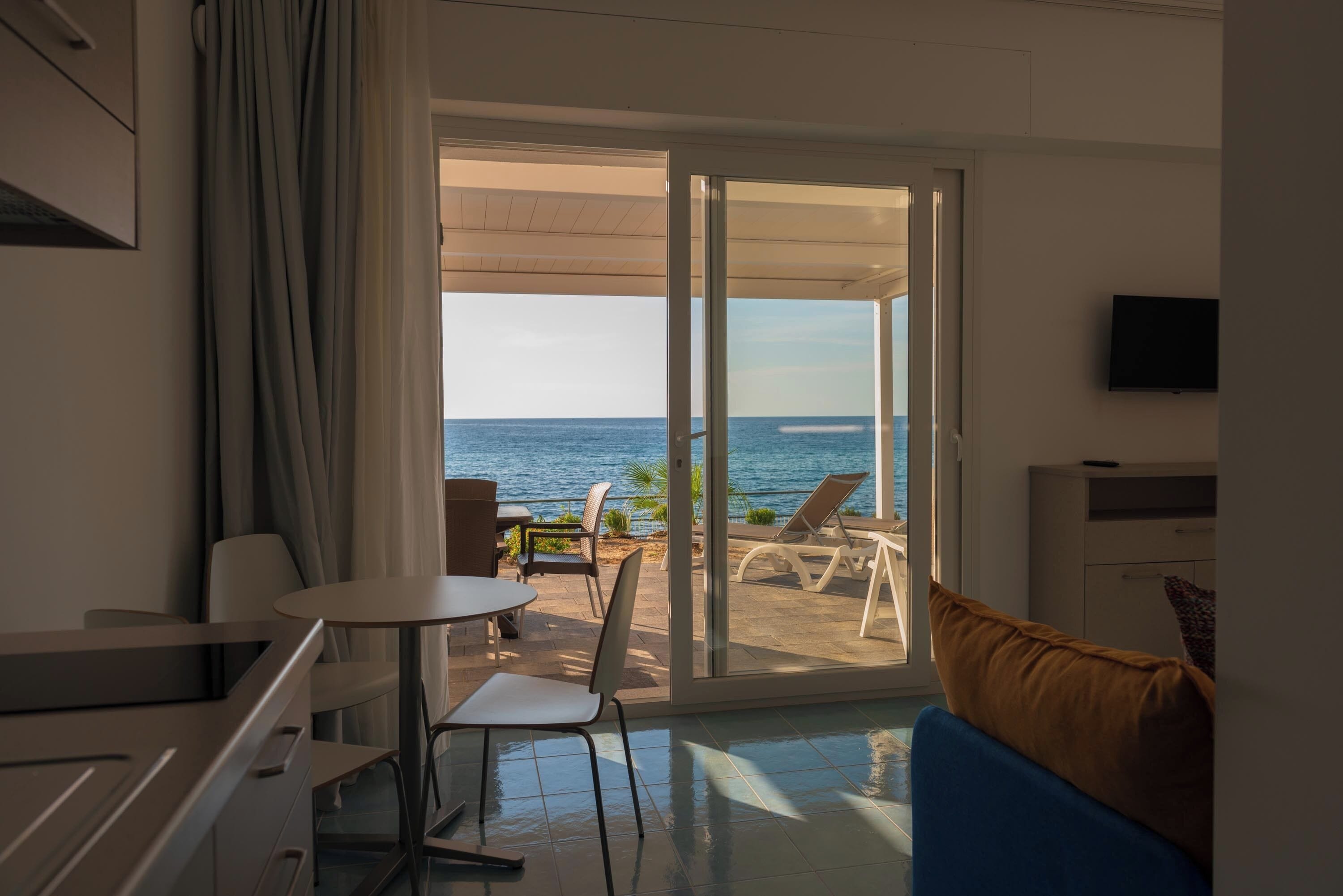 A room with a view of the sea, a dining area, and a TV on the wall.