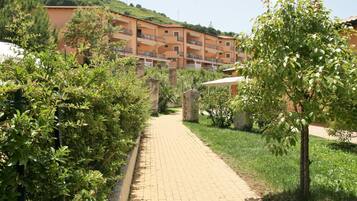 Un sendero pavimentado que conduce a un edificio de varios pisos con balcones, rodeado de follaje y construcciones de piedra.