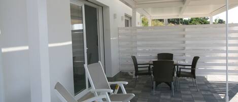 Un patio cubierto con sillas y una mesa blancas, una cerca blanca y una puerta que da a un corredor cubierto.