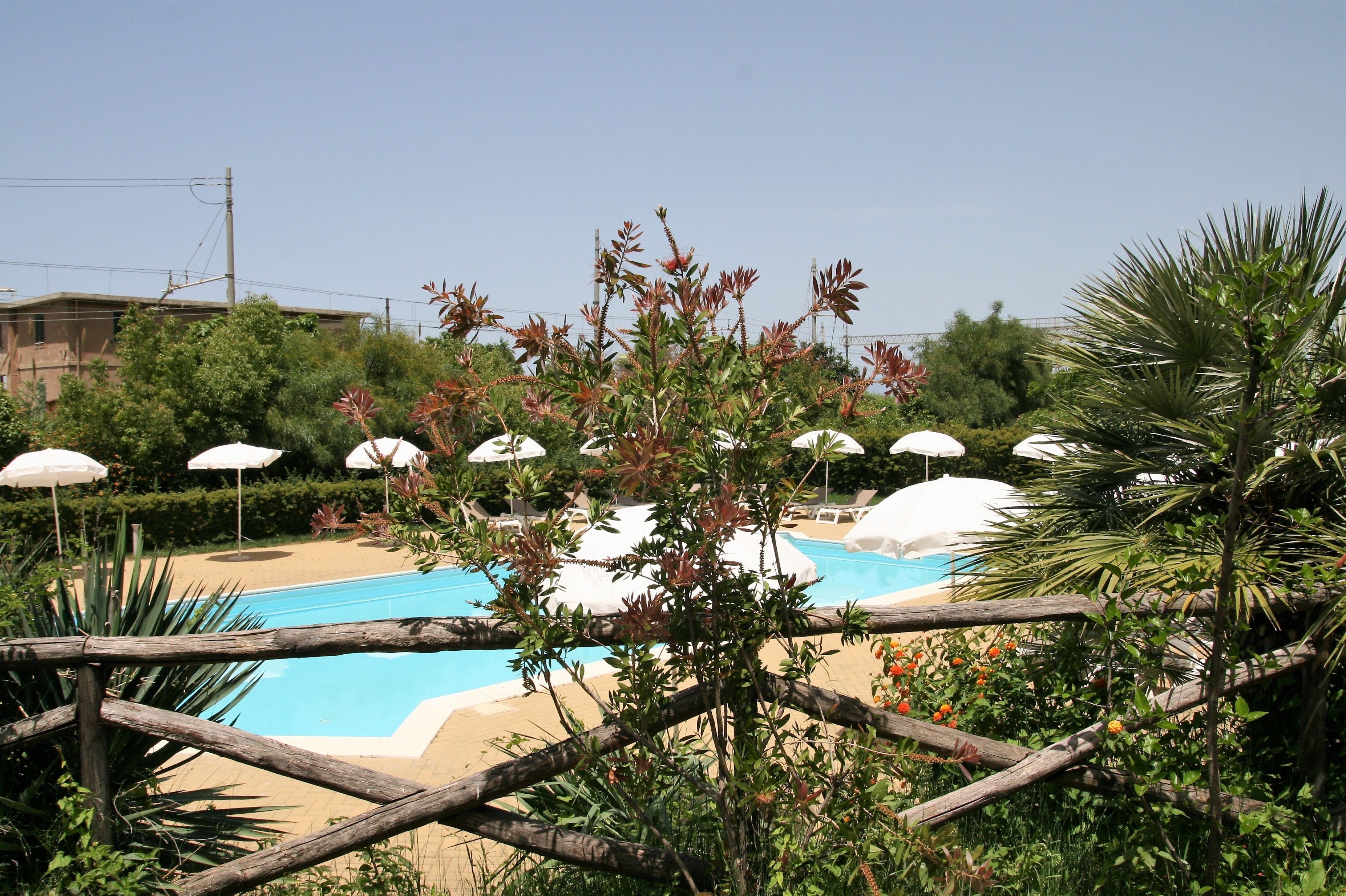A swimming pool with white umbrellas, surrounded by greenery and a wooden fence.
