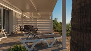 A covered outdoor seating area with lounge chairs and a table, a white building with a sliding door, and a clear sky.