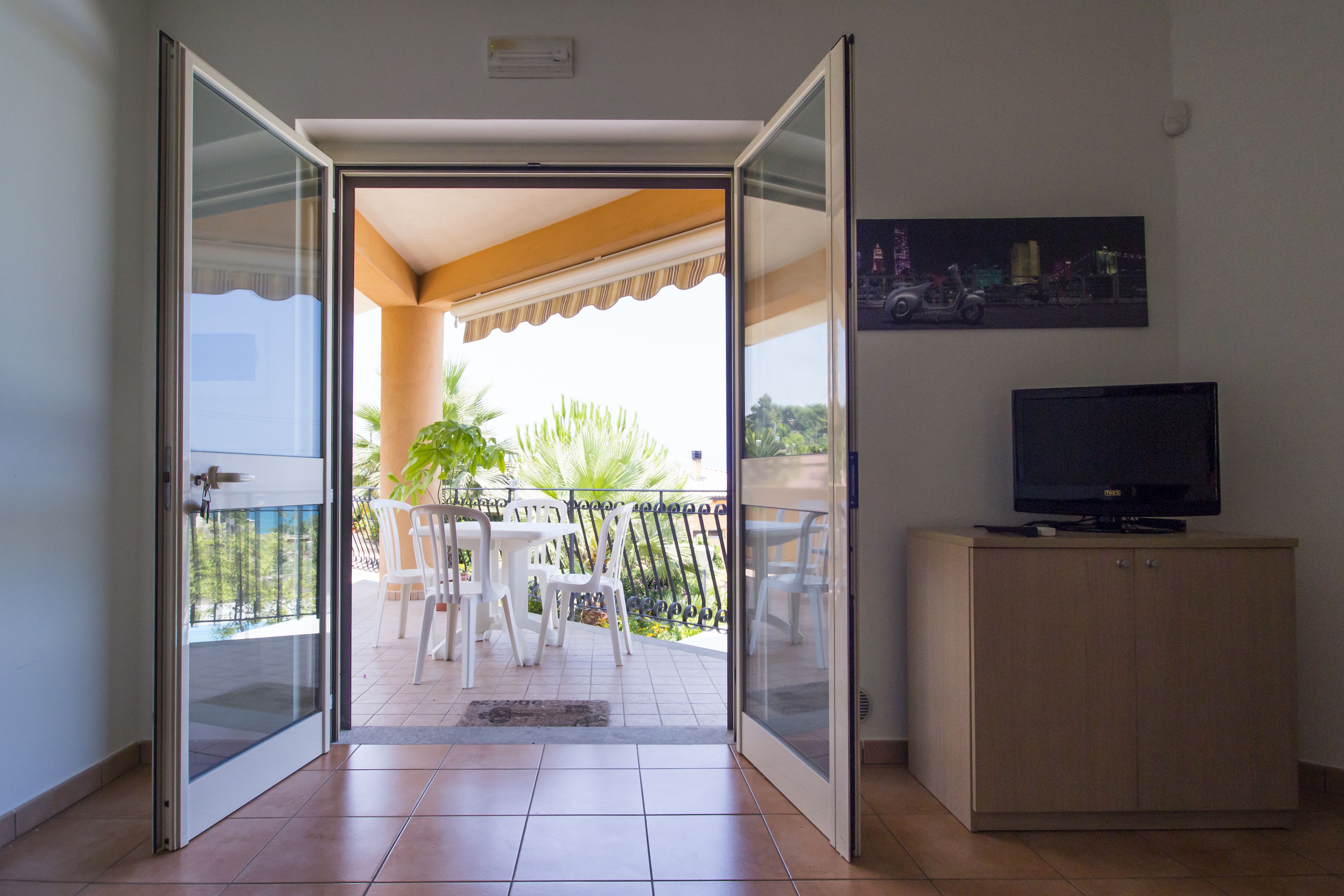 Une porte-fenêtre coulissante donne sur un balcon équipé d’une table et de chaises, ainsi que d’un téléviseur posé sur un meuble. On peut admirer une vue sur la ville depuis le balcon.