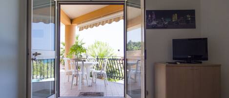 Une porte-fenêtre coulissante donne sur un balcon équipé d’une table et de chaises, ainsi que d’un téléviseur posé sur un meuble. On peut admirer une vue sur la ville depuis le balcon.