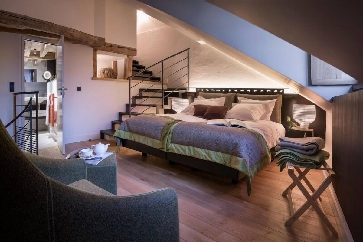 Une chambre avec un plafond en pente, un lit doté d’une tête de lit, un escalier et un petit coin salon.