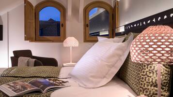A bedroom with a bed, a reading lamp, a bedside table with a cup and teapot, and a window with a view of buildings.