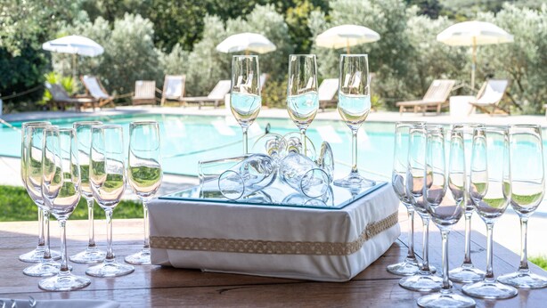 Mesa junto a la piscina con varios vasos de vino y una bandeja de copas, rodeada de sillones de descanso y sombrillas.