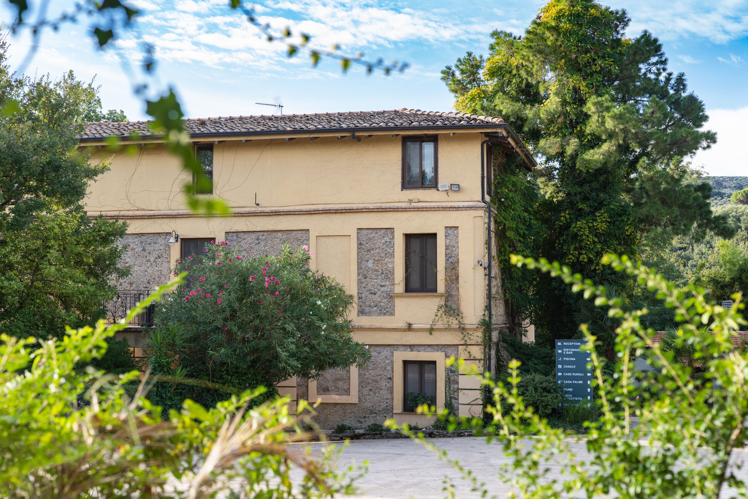 A two-story building with a stone facade and a tiled roof, surrounded by greenery and trees.