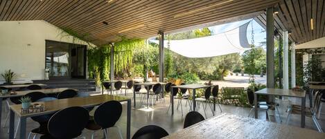 An outdoor dining area with tables and chairs, a wooden ceiling, and a view of the street and greenery.