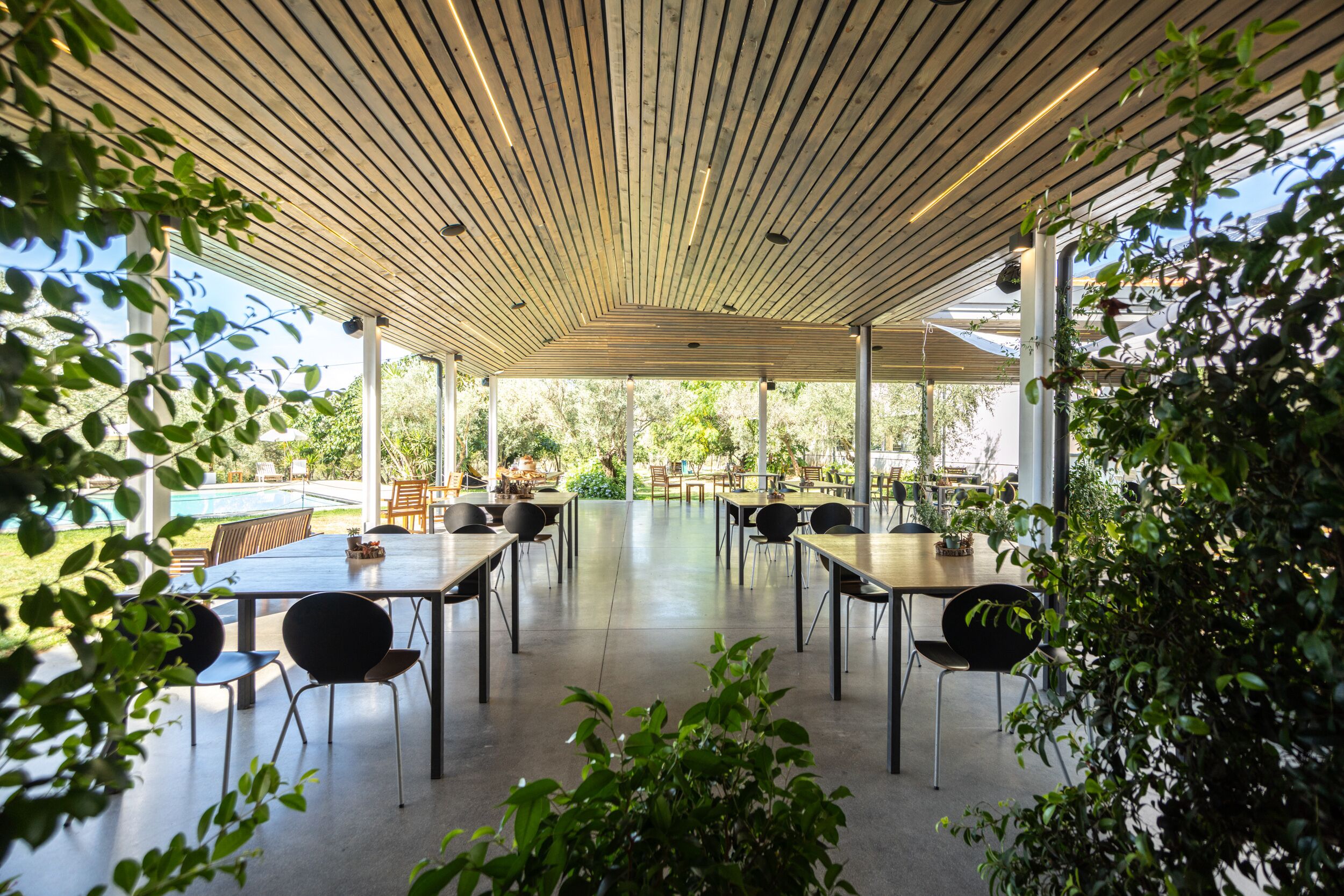 Un espacio comedor techado al aire libre, con mesas y sillas, rodeado de follaje y ventanas grandes.