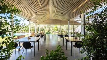 Un espacio comedor techado al aire libre, con mesas y sillas, rodeado de follaje y ventanas grandes.