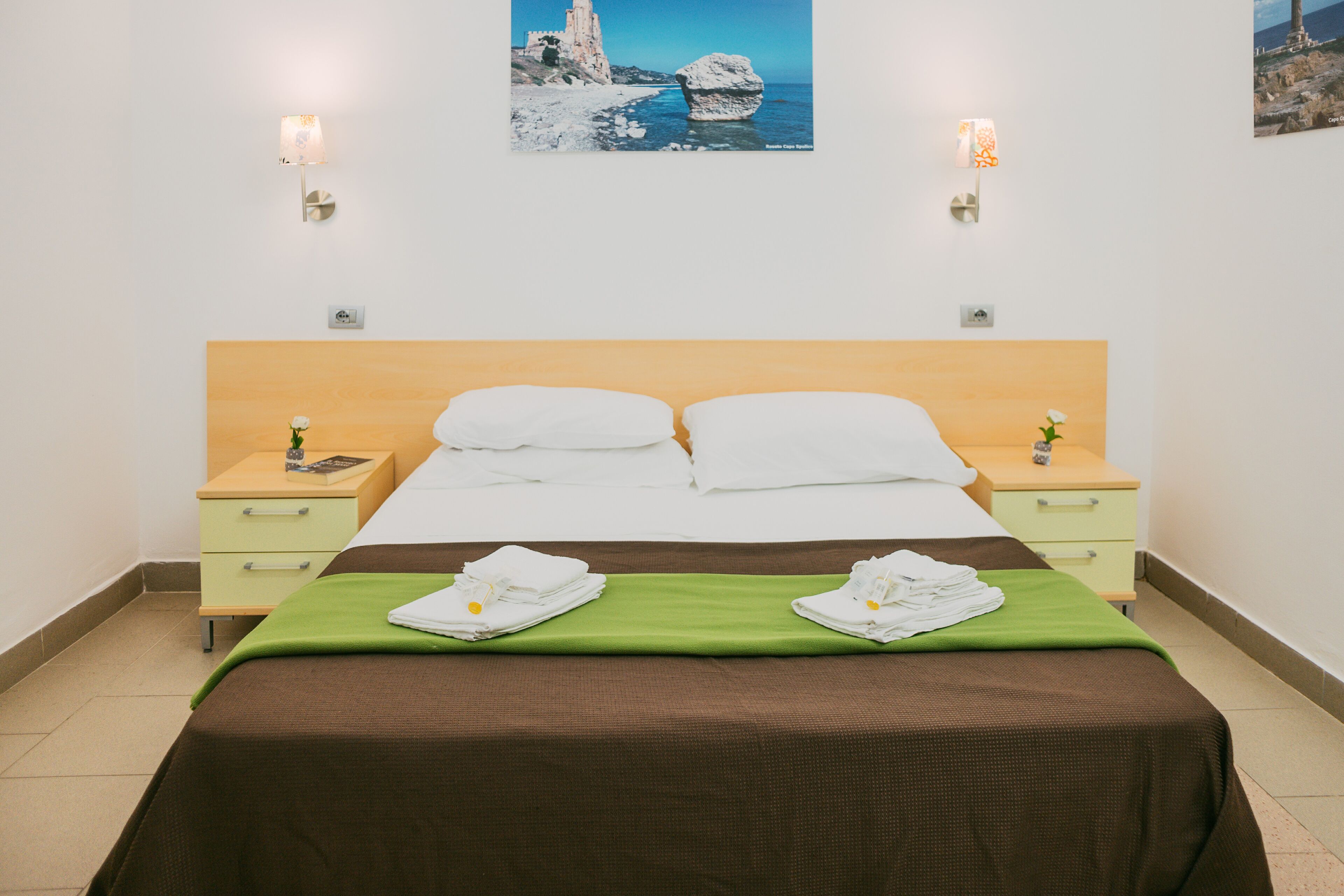 A neatly made bed with a brown bedspread, two white pillows, and a green blanket. There are two bedside tables with lamps, and a framed picture of a coastal scene is on the wall.